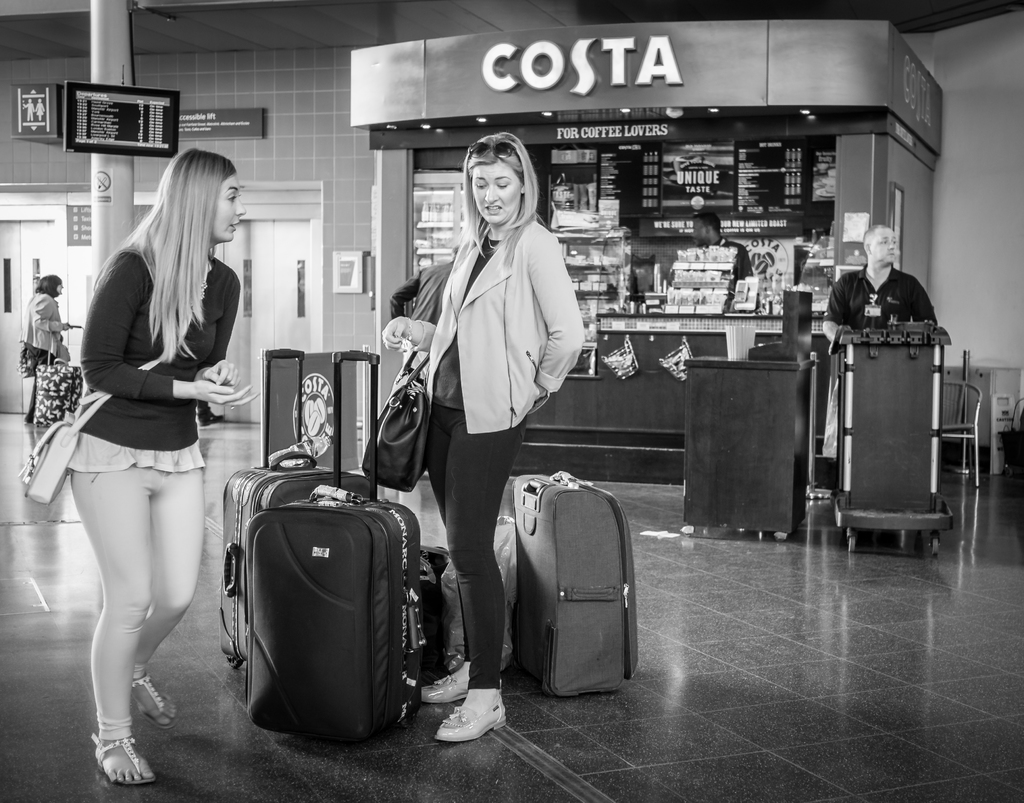 Zwei Frauen mit Gepäckstücken vor einem Kaffee-Shop an einem Ort, der wie ein Flughafen aussieht.