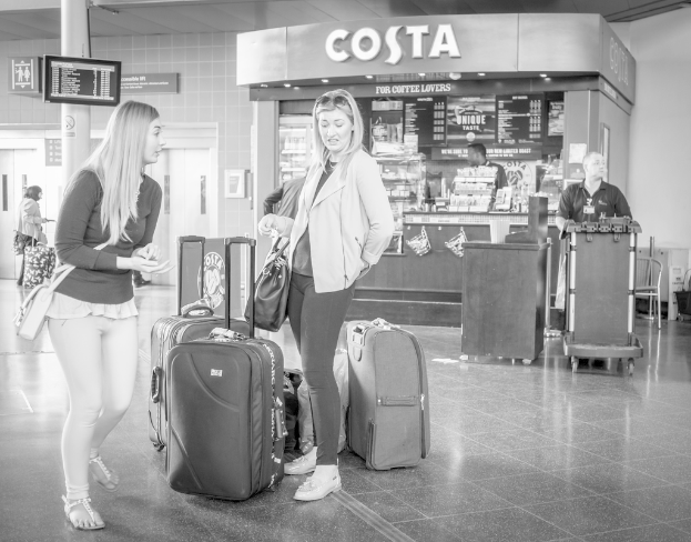 Zwei Frauen mit Gepäckstücken vor einem Kaffee-Shop an einem Ort, der wie ein Flughafen aussieht.