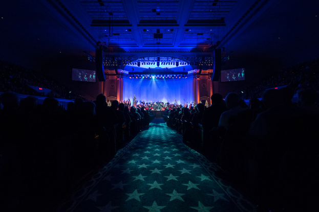 Ein großer Zuschauerraum mit Sitzenden, eine Bühne mit Musikern und eine Decke mit Lichtern und Fernsehern, was auf einen Konzertsaal für die Nationale Convention 2019 hindeutet.