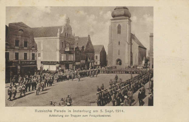 Ein Schwarz-Weiß-Foto eines Umzugs in Insterburg am 5. September 1914 mit vielen Menschen, Gebäuden und einem bewölkten Himmel sowie einem Text unten.