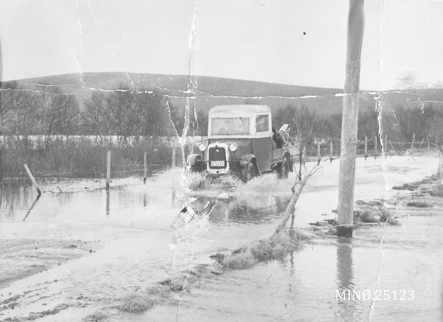 Alter Lkw fährt durch eine überflutete Straße mit Wasser drumherum, Polen, Bäume und einen Hügel im Hintergrund, unter einem sichtbaren Himmel, mit einem Wasserzeichen in der rechten unteren Ecke.