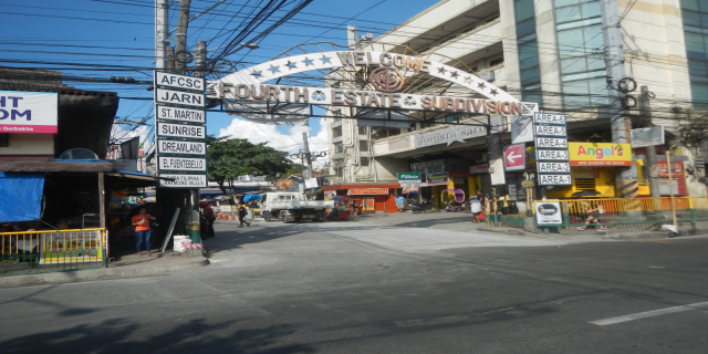 Eine belebte Stadtstraße mit Fahrzeugen, Fußgängern, Gebäuden und einem Schild mit der Aufschrift "Vierte-Estate-Unterteilung" in der Mitte, unter einem bewölkten Himmel.