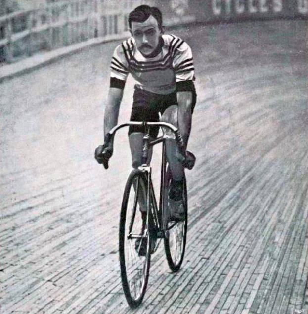 Ein Schwarz-Weiß-Foto eines Mannes, der auf einer Bahn ein Fahrrad fährt, mit einem Zaun und einer Tafel im Hintergrund, der als Sir Ernest Hemingway identifiziert wird.