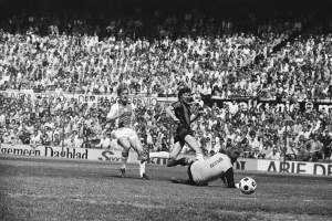 Schwarzes und weißes Foto von Männern, die auf einem Feld Fücball spielen, mit Zuschauern im Hintergrund und einem Banner 'Bundesliga 1978-79 Ajax 1-1 Hannover 96'.