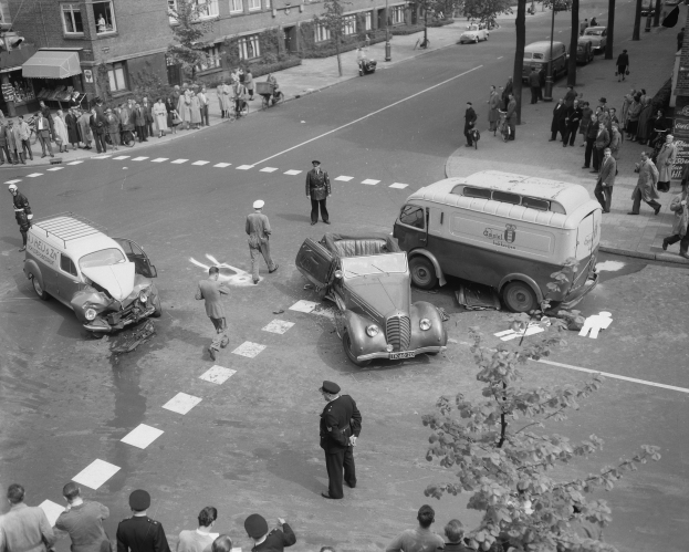 Schwarz-weißes Bild eines Autounfalls an einer Straßenkreuzung mit mehreren Fahrzeugen, darunter ein Lieferwagen, und einer Gruppe von Schaulustigen; Gebäude, Bäume, Strommasten und Texttafeln sind im Hintergrund zu sehen.