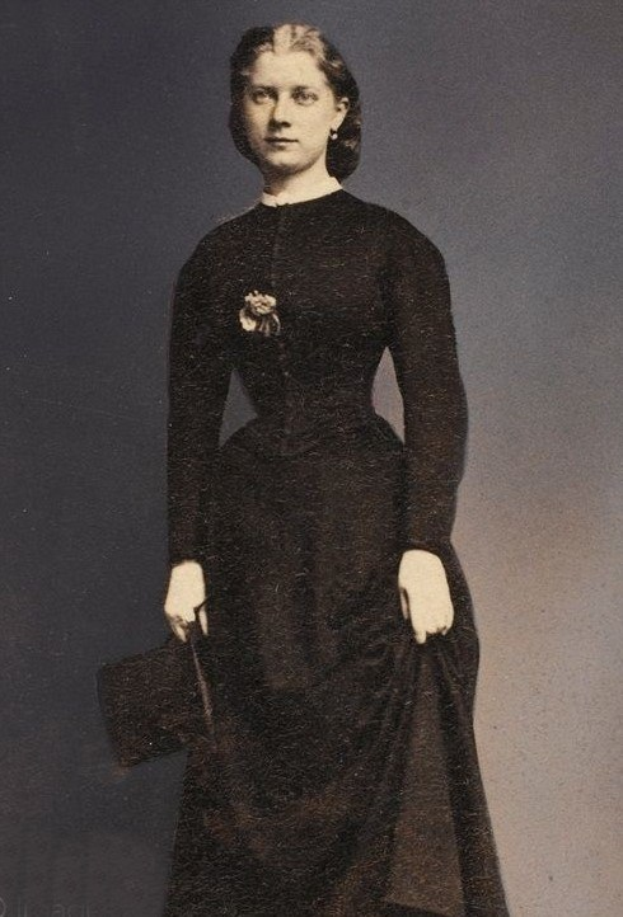 Ein Schwarz-Weiß-Foto einer älteren Frau in einem langen schwarzen Kleid mit hohem Kragen und langen Ärmeln, die mit ernster Miene steht, eine Tasche hält und ihr Haar zu einem ordentlichen Dutt frisiert hat.