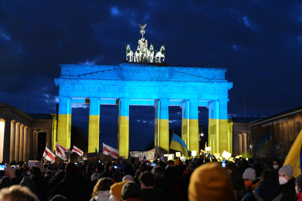 Eine Menge steht vor dem Brandenburger Tor in Berlin, hält Fahnen und Schilder, mit einem Banner auf der rechten Seite mit protestbezogenem Text.