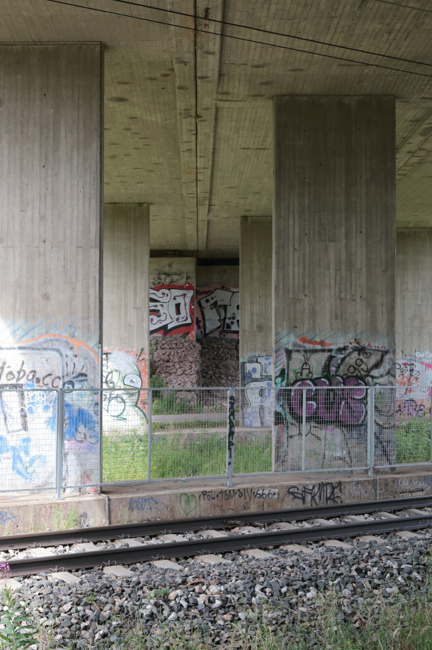 Eisenbahnschiene mit Graffiti an einer Betonwand, umgeben von Steinen, Gras, einem Zaun, tragenden Säulen, Oberleitungen und einem Dach.