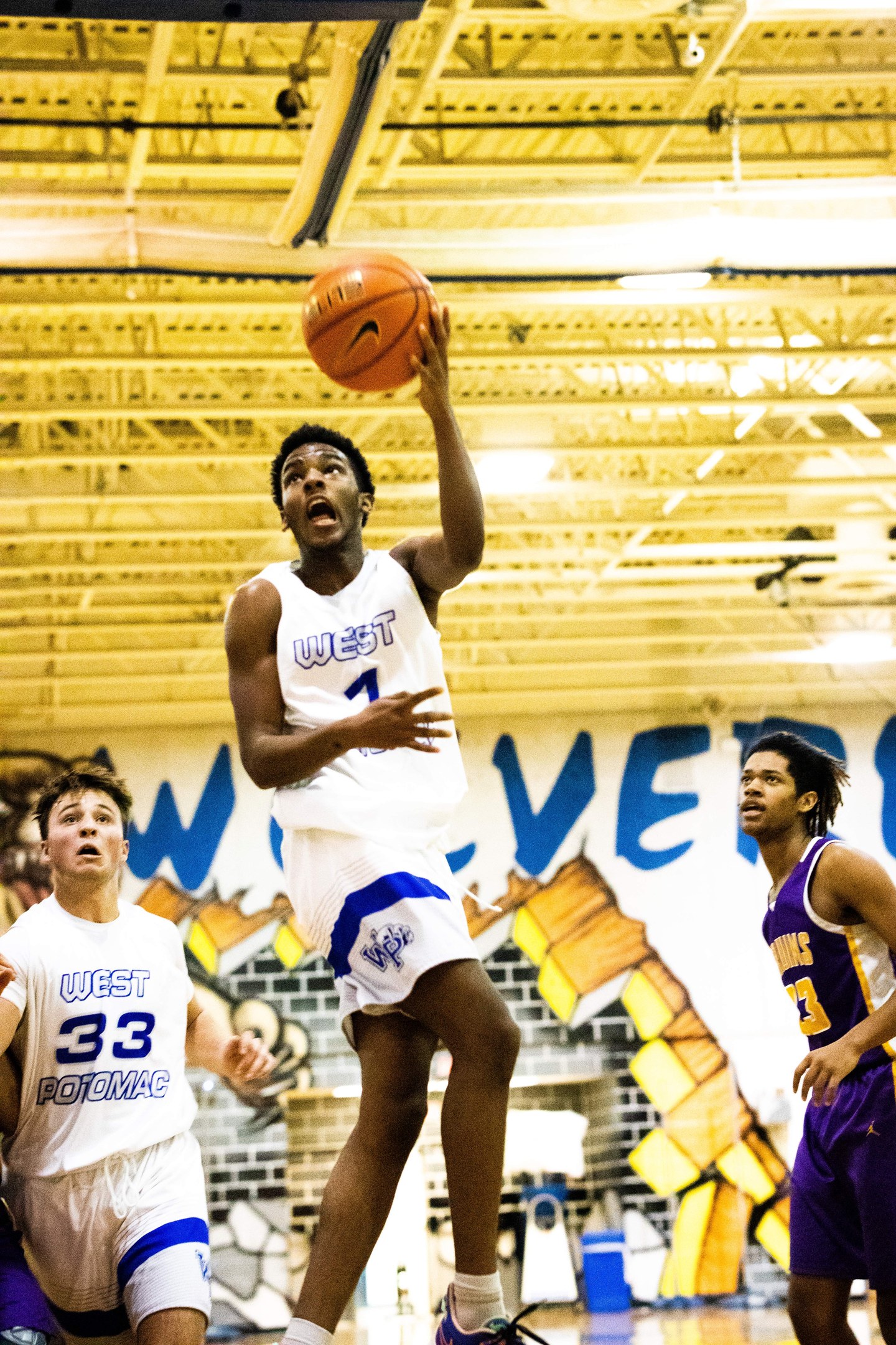 Junge Männer spielen Basketball in einer Turnhalle mit einer bemalten Wand, Deckenlampen und einem Spieler mit Turnschuhen.