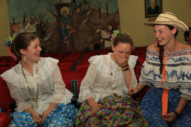 Drei Frauen in weißen Oberteilen und Röcken sitzen und lachen, mit roten Stühlen und Wandkunst im Hintergrund.