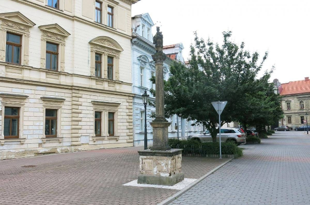 Eine Statue auf einem Sockel in einem Stadtplatz mit umgebenden Gebäuden, Straßeninfrastruktur, Kraftfahrzeugen, Vegetation und einem klaren Himmel.