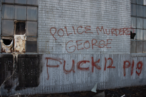 Ein Gebäude mit Graffiti, das "Polizei Mord George Fuck 12 P19" auf der Seite trägt, mit Fenstern und diversen Gegenständen vorne auf dem Boden.