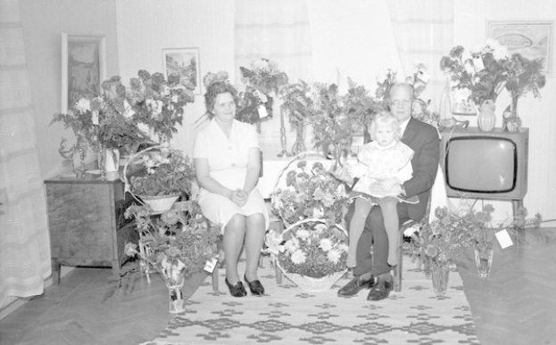 Schwarzes und weißes Foto einer Familie aus drei Personen - einem Mann, einer Frau und einem Kind - die auf einem Sofa vor einem Fernseher sitzen, mit Blumenvasen auf den Seitentischen, einem Teppich auf dem Boden, Vorhängen im Hintergrund und Fotorahmen an der Wand.