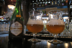 Eine Flasche mit Logo und zwei Weingläser auf einem Tisch, mit vielen Gläsern, Flaschen und Regalen im Hintergrund.