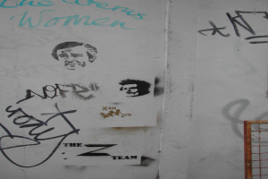 Ein verrosteter Zaun steht vor einer Wand mit Zeichnungen und Wörtern.