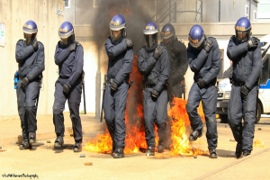 Menschen in Helmen stehen vor einem Feuer mit verschiedenen Gegenständen auf dem Boden, Gebäuden im Hintergrund, einem Fahrzeug und einem Plakat und einer Tafel an der linken Wand; Text ist am unteren Bildrand sichtbar.