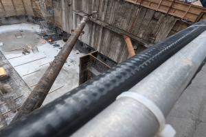 Eine Baustelle mit einem großen Metallrohr in der Mitte, umgeben von Arbeitern, Holzgegenständen und diverser Ausrüstung, mit Fahrzeugen und einem Gebäude im Hintergrund.