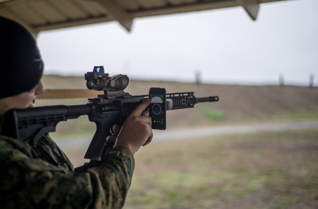 Ein Mann hält eine Waffe und bedient ein Mobiltelefon im Vordergrund, mit einem Schuppen darüber und einer Grasfläche, Straße, Polen und Himmel im Hintergrund.