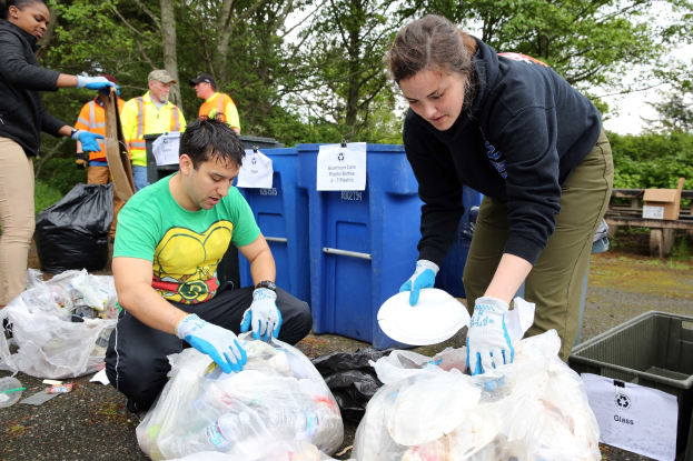 Eine Gruppe von Menschen mit Handschuhen sammelt Müll in einem Park, umgeben von Plastikabfällen, Flaschen und anderen Gegenständen, mit einem Müllcontainer und einer Holzbank rechts und Bäumen und einem blauen Himmel im Hintergrund.