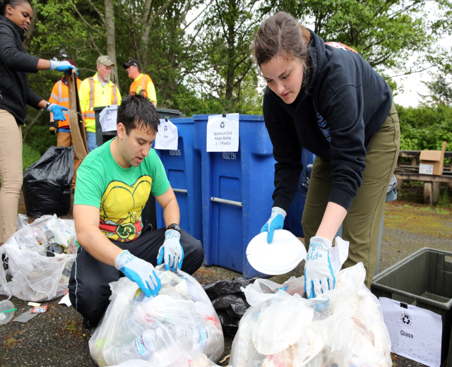 Eine Gruppe von Menschen mit Handschuhen sammelt Müll in einem Park, umgeben von Plastikabfällen, Flaschen und anderen Gegenständen, mit einem Müllcontainer und einer Holzbank rechts und Bäumen und einem blauen Himmel im Hintergrund.