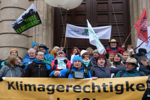 Eine Gruppe von Menschen steht vor einem Gebäude mit Säulen und einer Tür, hält ein Transparent mit Text, Fahnenstangen und Luftballons und nimmt an einem Klimastreik in Deutschland teil.