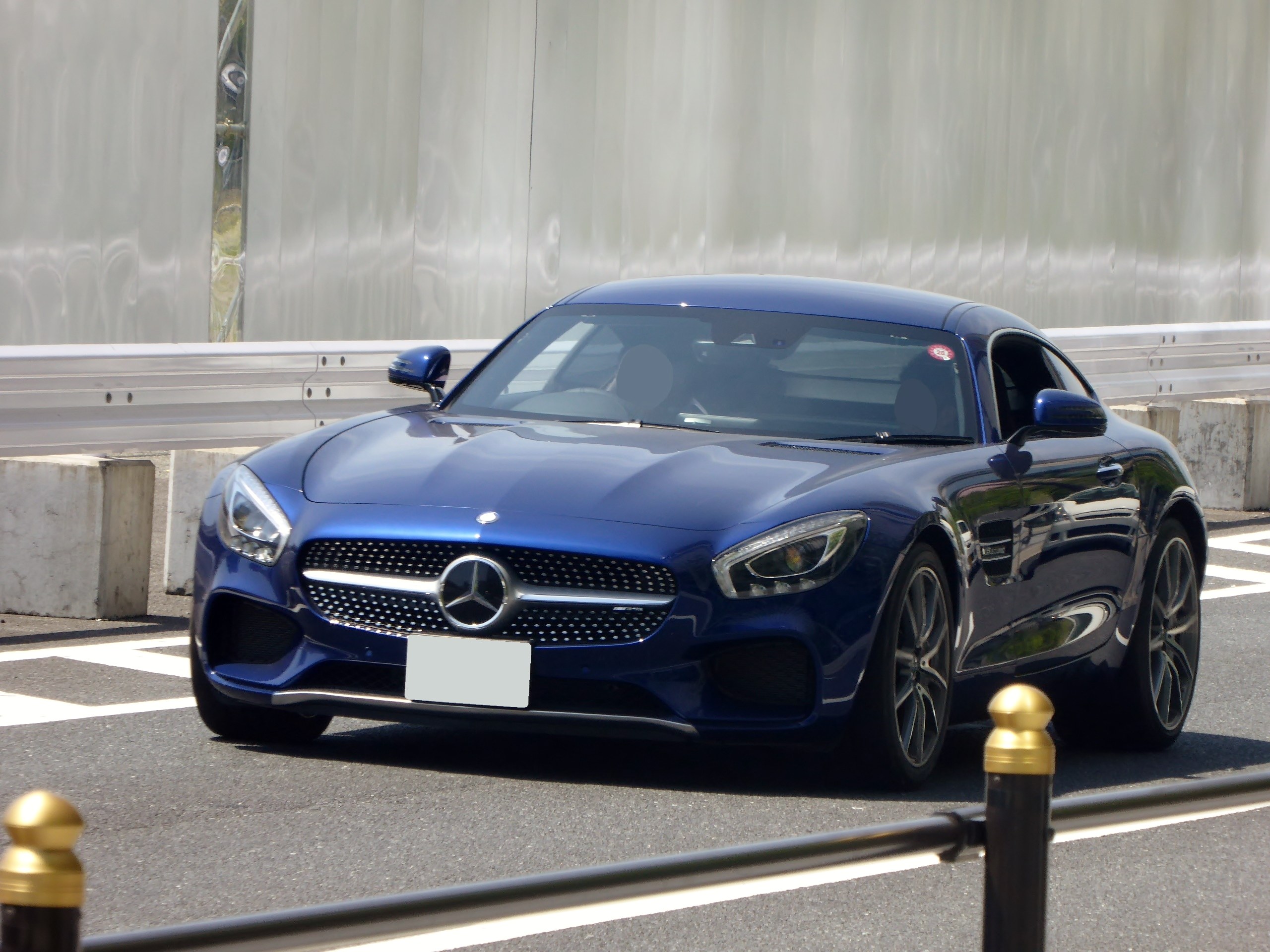 Ein blauer Mercedes-Benz AMG GT Coup√© mit sportlicher, aerodynamischer Gestalt, eckigen Scheinwerfern, großem Kühlergrill und glänzenden Rädern fährt auf einer Straße mit einer Wand im Hintergrund.