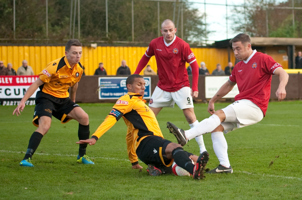 Spieler in blauen und roten Uniformen spielen ein Spiel auf einem Rasenfeld mit einem Ball, während Zuschauer außerhalb des Spielfelds stehen und sie anfeuern, mit einem Baum und Himmel im Hintergrund.