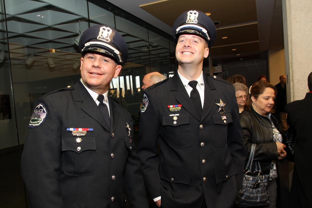 Zwei lächelnde Beamte im Vordergrund, mit Menschen in der Mitte, Glasfenster links, Lampen und Decke oben und Säulen und Menschen rechts.