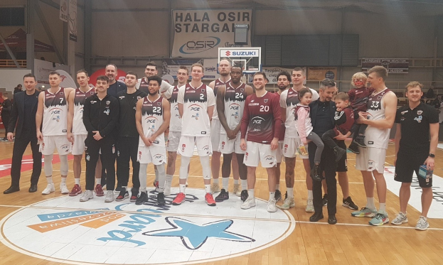 Männer- und Kindergruppe auf einem Basketballfeld mit Schildern "Hala Osir Starga", sitzende Zuschauer, ein Basketballkorb, Metallstangen, Deckenbeleuchtung und eine Wand im Hintergrund.