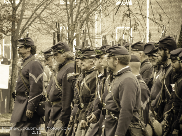 Mehrere Männer mit Mützen auf der Straße, einige halten Waffen, mit Bäumen und einem Gebäude im Hintergrund; ein Wasserzeichen ist in der linken unteren Ecke sichtbar, schwarz-weißes Bild.