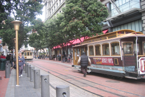 Ein Zug mit Schriftzug fahrt auf Schienen, die in eine Stra├če eingebettet sind, vorbei an einer Person, kleinen Pfosten mit Seilen links, Stra├čenlaternen, B├Ąumen, vielen Menschen, einem M├╝lleimer links und Geb├Ąuden mit Leitern rechts.