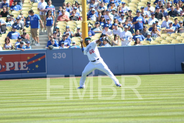 Ein Mann in einer Mütze und Sonnenbrille versucht, einen Ball zu werfen, auf einem grünen Feld mit einer Zuschauermenge, die auf Stühlen sitzt und steht, einige tragen ebenfalls Mützen, und ein Pfahl ist im Hintergrund zu sehen.