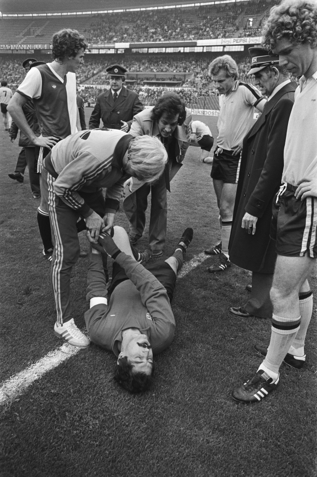 Schwarzes und weißes Bild eines Fußballspielers auf dem Boden umgeben von einer Gruppe von Männern, mit einer Menschenmenge im Hintergrund.