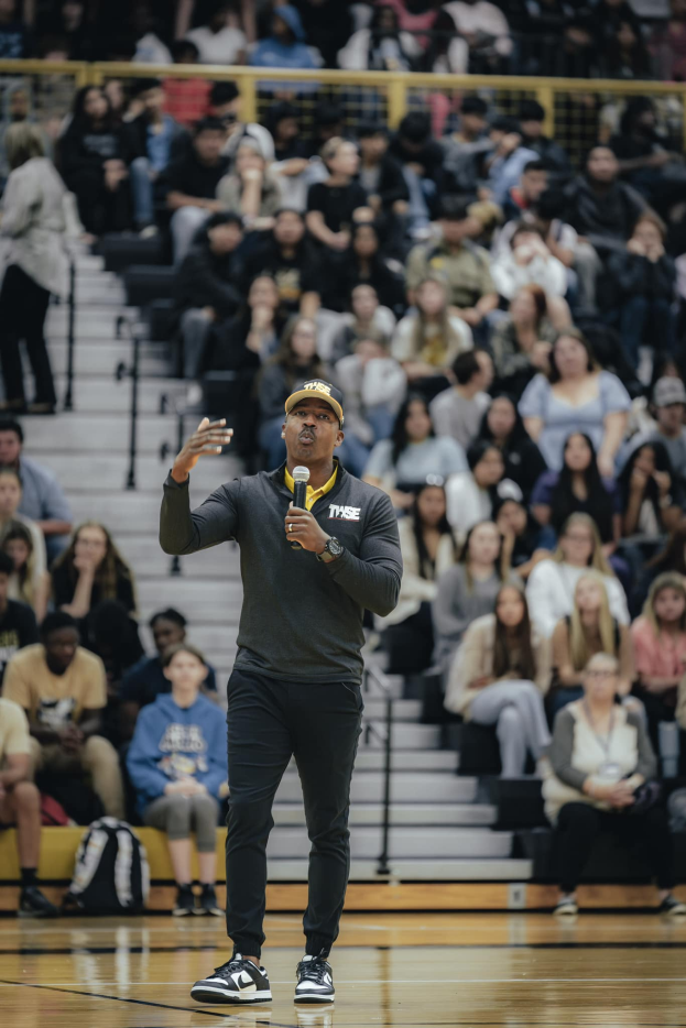Ein Mann mit Mütze hält ein Mikrofon auf einem Basketballfeld, mit einer Gruppe von Menschen auf einer Treppe und Taschen auf dem Boden dahinter.