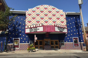 Das Esquire Theatre, ein blaues Gebüude mit einem bunten Wandgemülde, umgeben von Büschen, Laternen, Strommasten mit Drühten und einer Person auf dem Gehweg, mit Türen, Fenstern und Schildern mit Text.