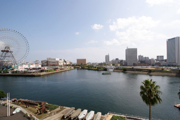 Eine Stadtansicht mit Wolkenkratzern, einem Riesenrad, Vergnügungsanlagen, Schiffen auf dem Wasser und verschiedenen Strukturen wie Geländern, Skulpturen und Barrieren, alles unter einem bewölkten Himmel mit Bäumen und Büschen im Vordergrund.