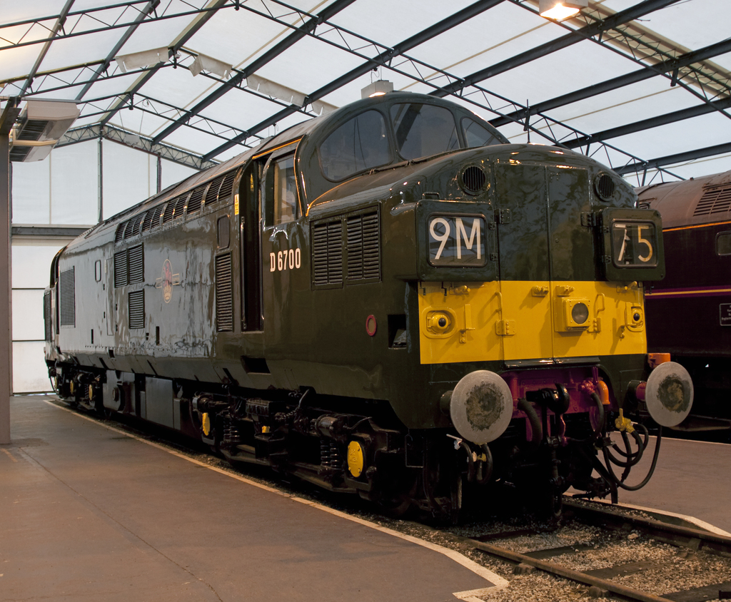 Ein Zug steht auf den Schienen neben einem Bahnsteig mit einem Mast auf dem Dach, der Metallstangen mit Lampen trägt.