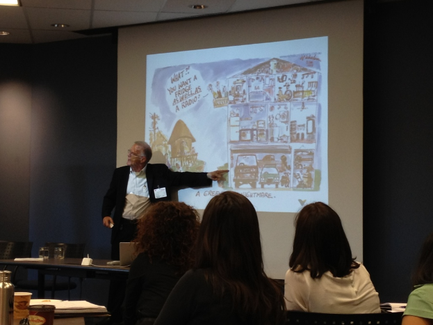 Ein Mann in weißem Hemd und schwarzer Jacke zeigt auf ein auf einem Bildschirm angezeigtes Fahrzeug in einem Konferenzsaal, wobei drei Frauen im Vordergrund und ein Tisch mit einer Wasserflasche, einem Glas und einem Buch links zu sehen sind.