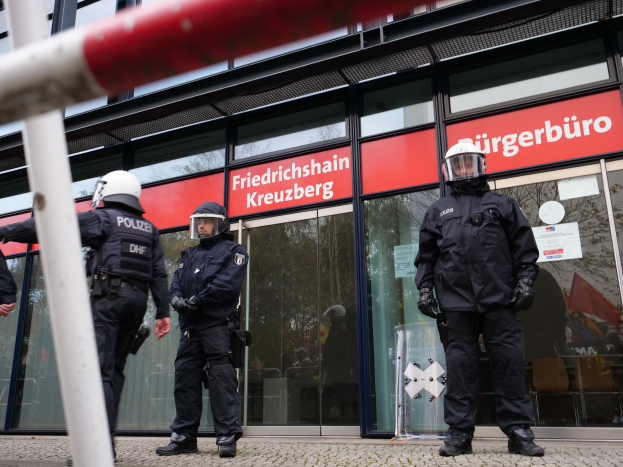 Eine Gruppe von Polizisten in Helmen und Handschuhen steht vor einem Glaswandgebäude mit angeklebten Papieren, neben einem Pfahl, mit "Friedrichshain Kreuzberg" Schildern im Hintergrund.
