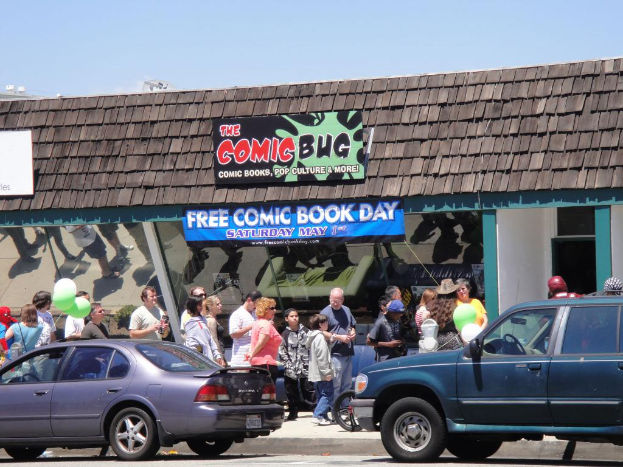 Eine Gruppe von Menschen vor einem Gebäude mit Glastüren und einem Dach, zwei Autos auf der Straße, grüne Luftballons, ein Banner und ein Namensschild auf dem Dach unter einem klaren Himmel.