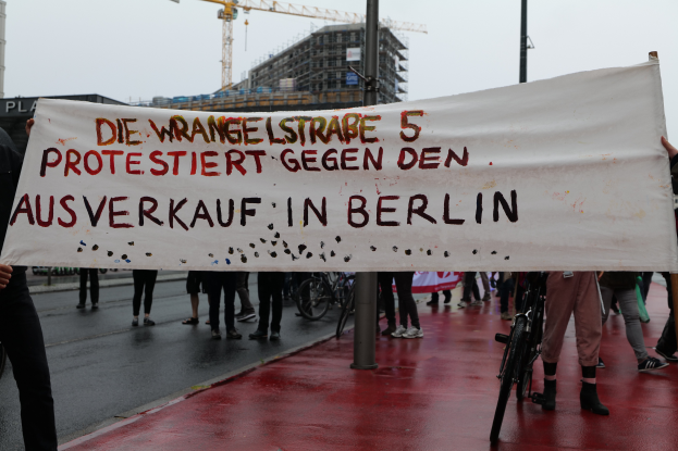 Eine Gruppe von Menschen steht auf einer Straße und hält ein Banner in der Hand, auf dem "Die Wrangellstraße 5 protestiert gegen den Ausverkauf in Berlin" steht. Im Hintergrund sind Fahrräder, Stangen, Gebäude und ein Kran zu sehen, über ihnen der Himmel.