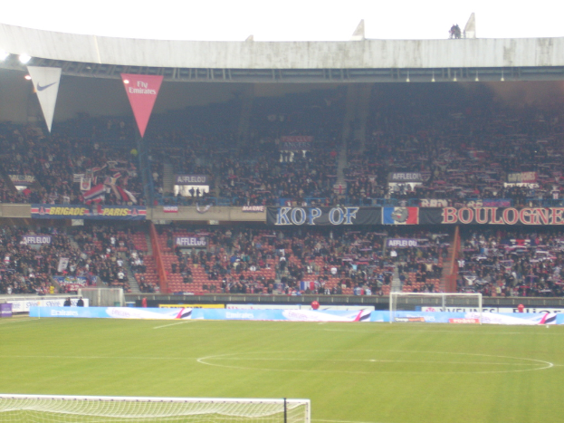 Ein Rasenboden mit Torstangen auf beiden Seiten, Zuschauern im Hintergrund und zahlreichen Werbetafeln in der Mitte und darüber.