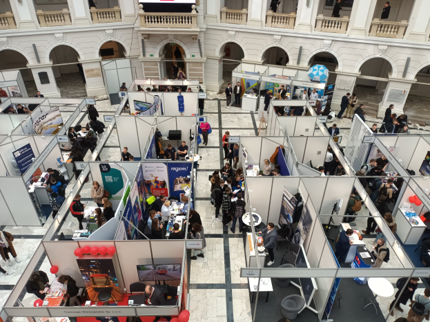 Große Besuchermenge auf einer internationalen Fachmesse mit Ständen, Bannern, Tischen, Stühlen und Ballons in einem Raum mit Architektur Bögen, Säulen, Geländern und einem Bildschirm im Hintergrund.