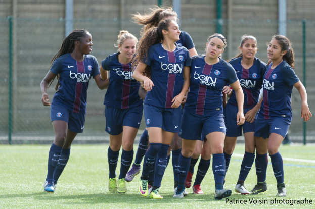 Gruppe junger Frauen beim Fußballspielen auf einem Rasenplatz mit Maschendrahtzaun und einer Wand im Hintergrund, Text in der rechten unteren Ecke lautet "Paris Saint-Germain Women's Soccer".