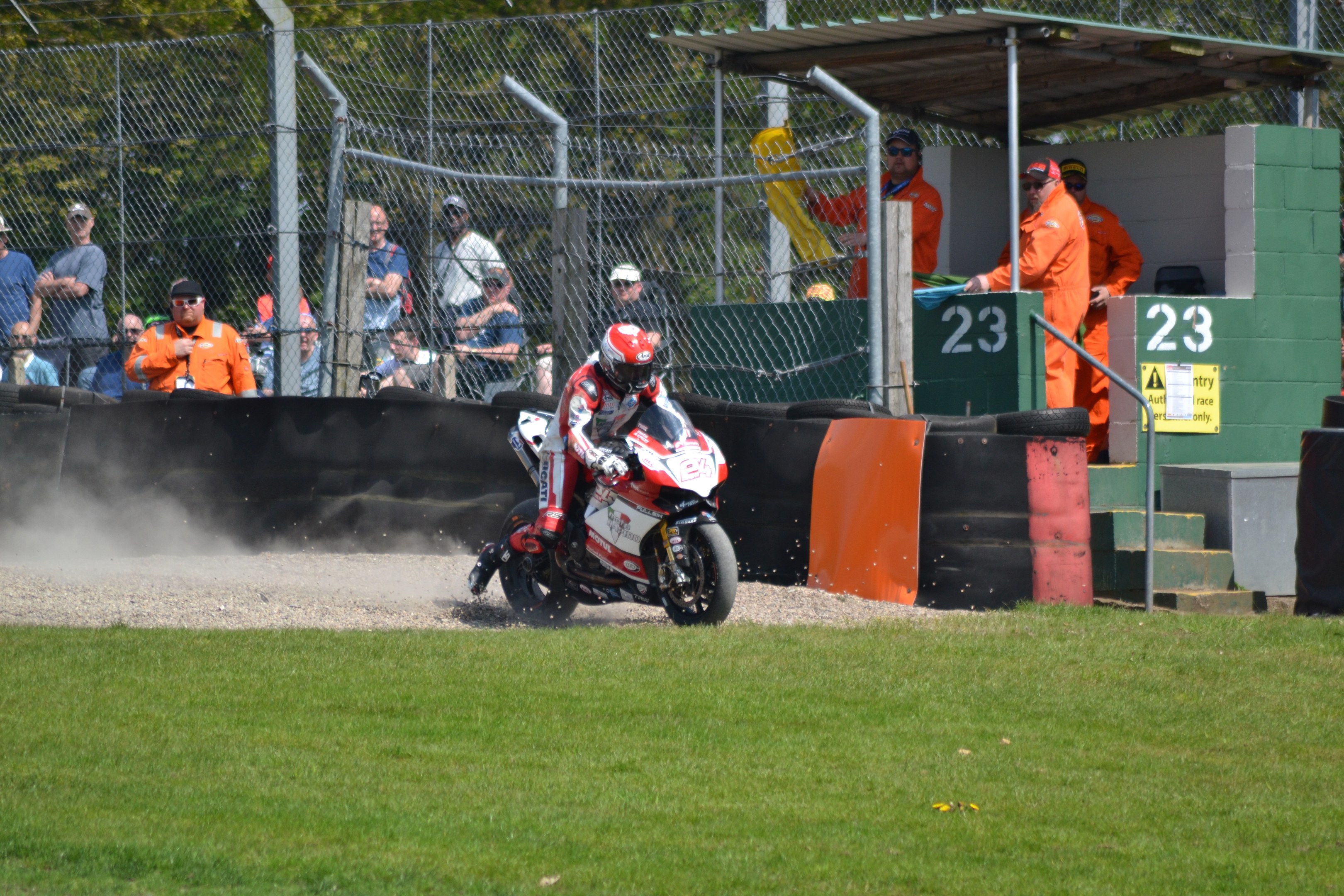 Eine Person in Helm und Handschuhen auf einem Motorrad auf einem grasbewachsenen Feld, mit Zuschauern, Absperrung, Treppe und einem Schuppen im Hintergrund.