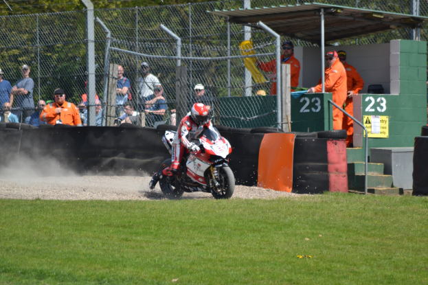 Eine Person in Helm und Handschuhen auf einem Motorrad auf einem grasbewachsenen Feld, mit Zuschauern, Absperrung, Treppe und einem Schuppen im Hintergrund.