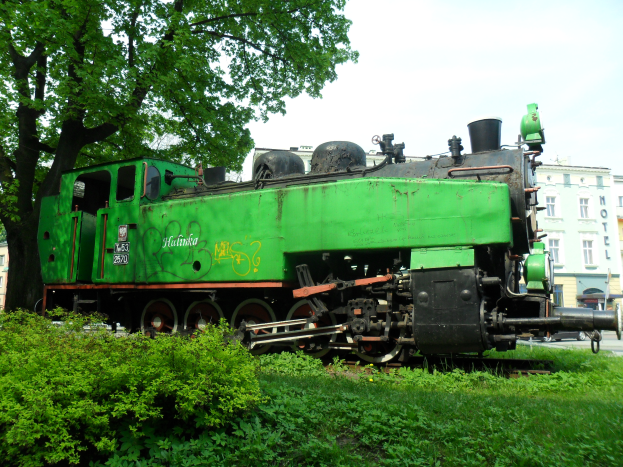 Ein grüner Zug auf einem grünen Feld mit Gebäuden, Fahrzeugen und einem klaren blauen Himmel im Hintergrund.