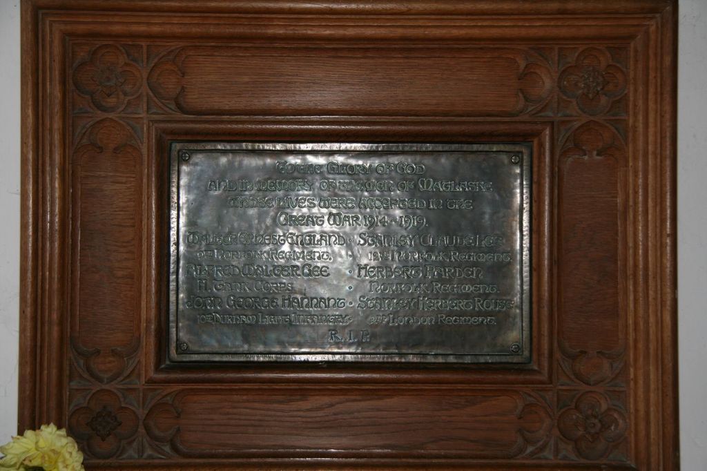 Eine Gedenktafel mit Text ist an einer braunen Wand oder Holztür angebracht, mit einem gelben Blumentopf in der linken unteren Ecke.