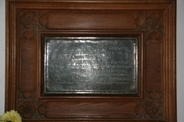 Eine Gedenktafel mit Text ist an einer braunen Wand oder Holztür angebracht, mit einem gelben Blumentopf in der linken unteren Ecke.