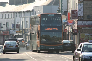 Eine Straße mit Autos und einem Bus vor Gebäuden mit Wänden, Fenstern, Tellern und Dächern, mit Plakaten und Bannern an den Wänden und einem Pfosten mit einer Straßenlaterne.